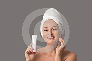 Portrait of beautiful mature woman with perfect skin holding tube of cream on background