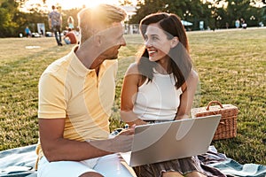 Portrait of beautiful middle-aged couple smiling while using laptop computer during picnic in summer park