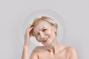 Portrait of beautiful middle aged woman smiling at camera, adjusting her hair, posing isolated against grey background