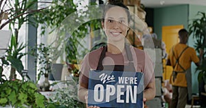 Portrait of beautiful Middle Eastern lady in apron standing in flower store holding Yes we are open sign