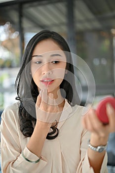 Portrait of a beautiful millennial Asian woman applying red lipstick, putting on her makeup