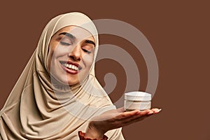 Portrait of beautiful modern muslim woman with natural make-up dressed in beige hijab smiling with closed eyes and holding cream