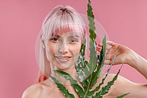 Portrait of beautiful naked woman with pink hair smiling at camera, posing with plant leaf isolated over pink studio