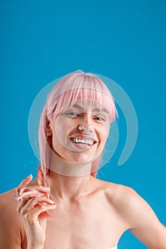 Portrait of beautiful naked woman smiling at camera, touching her pink straight hair while posing isolated over blue