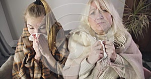 Portrait of beautiful old lady and young girl sitting on couch together and sneezing. Ill grandmother and granddaughter