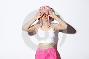 Portrait of beautiful party girl in pink wig, holding macaroons over eyes and pouting silly, standing over white
