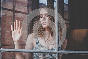 Portrait of beautiful pensive woman with curly hait and touching window glass