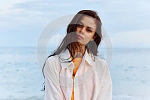 Portrait of a beautiful pensive woman with tanned skin in a white beach shirt with wet hair after swimming on the beach
