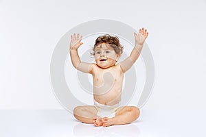 Portrait of beautiful playfull toddler boy, baby in diaper sitting with raised hands isolated over white studio