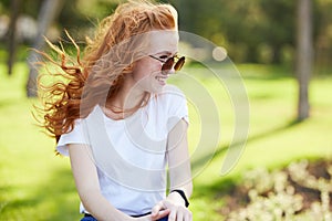 Portrait of a beautiful red-haired girl who sits in the park and looks away. The wind develops her hair and the girl smiles