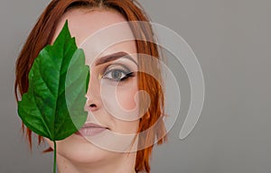 Portrait of a beautiful red-haired woman, half of her face closed by a large green leaf of a Chinese rose. Women`s health, natura