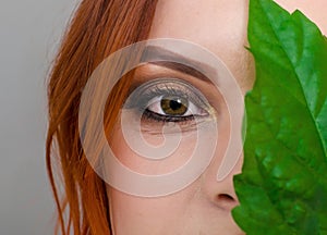 Portrait of a beautiful red-haired woman, half of her face closed by a large green leaf of a Chinese rose. Women`s health, natura