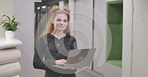 Portrait of beautiful redhead Caucasian woman standing with laptop in open space office, looking at camera and smiling