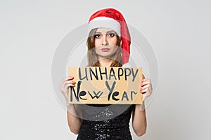 Portrait of a beautiful sad brunette woman in christmas hat and festive black dress holding a cardboard sign `unhappy new year ` o