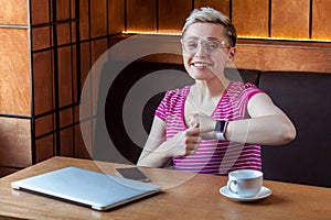 Portrait of beautiful satisfied happy young bussineswoman with blonde short hair in pink t-shirt is sitting in cafe, pointing