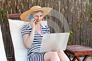 Portrait of beautiful satisfied young adult businesswoman in hat and dress sitting on sunbed with laptop and working online,