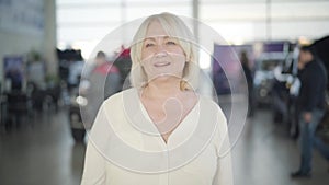 Portrait of beautiful senior blond Caucasian woman with grey eyes looking at camera and smiling. Mature lady standing in