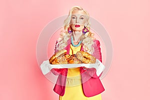 Portrait of beautiful senior woman in bright makeup and colorful clothes colding freshly baked buns against pink studio