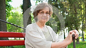 Portrait of beautiful senior woman with cane sitting on a bench in park, looking at camera and happily smiling