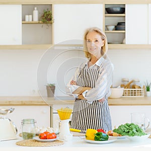 Portrait of beautiful senior woman standing in kitchen and smiling at camera while preparing healthy food