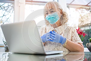 Portrait of beautiful senior woman wearing protective mask and gloves having video chat on laptop at home.