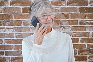 Portrait of beautiful senior woman white haired using phone standing against a brick wall smiling