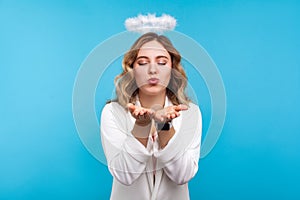 Portrait of beautiful sensual angelic woman with halo on wavy hair sending air kiss. indoor studio shot isolated on blue