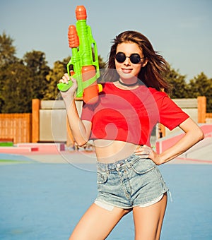 Portrait beautiful girl posing in denim shorts, white T-shirt and sunglasses in the skate park with water gun on a warm summe