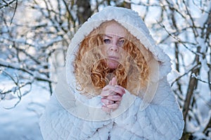 portrait of beautiful sexy mature redhead woman, with red curly hair in white hooded coat in winter nature sun backlight, snowy