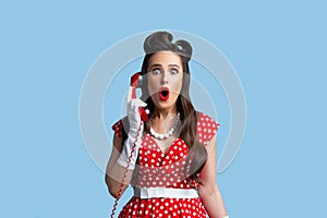 Portrait of beautiful shocked young pinup woman wearing retro dress, talking on landline phone on blue studio background