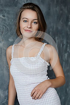 Portrait of beautiful slim woman in trendy white summer sarafan with smile leaning on gray wall, free copy space