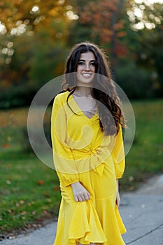 Portrait of a beautiful smiling teen girl with brown wavy hair, wear in yellow dress, posing in a park background.