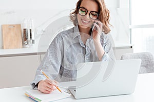 Portrait of beautiful smiling woman in glasses taking notes while speaking on mobile phone, indoors