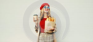 Portrait beautiful smiling woman holding coffee cup, grocery shopping paper bag with long white bread baguette over blank gray