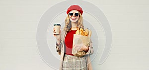 Portrait beautiful smiling woman holding coffee cup, grocery shopping paper bag with long white bread baguette over blank gray