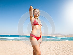 Portrait of beautiful smiling young woman with long hair wearing red bikini posing on the sea beach and looking in