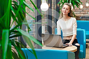 Portrait of beautiful smiling woman sitting on a comfortable chair in a cafe with black laptop. Pretty student doing work with lap