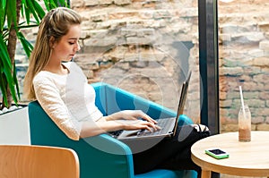 Portrait of beautiful smiling woman sitting on a comfortable chair in a cafe with black laptop. Pretty student doing work with lap