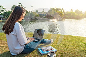 Portrait of beautiful smiling woman sitting on green grass in park with legs crossed during summer day and writing notes with pen