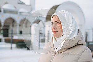 Portrait of beautiful smiling young Muslim woman in headscarf in light clothing against the background of mosque in the winter
