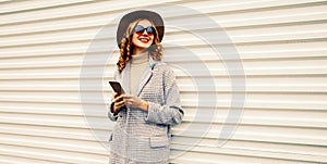 Portrait of beautiful smiling young woman with smartphone wearing coat jacket, hat on white background