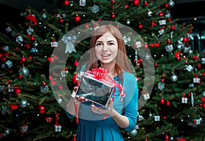 Portrait of beautiful smiling young woman, standing near Christmas tree, holding gift