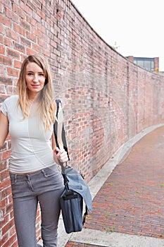 Twenty-something woman in white top jeans posing by red brick wall with messenger bag, copy space