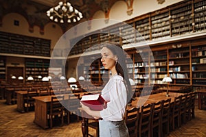 Portrait of a beautiful student in a white blouse studying in the university library, standing with a book in hand and looking