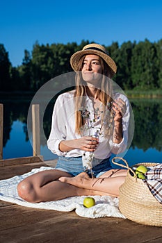 Portrait beautiful stylish romantic retro girl holding flower branch in hand while sitting at pier near the lake