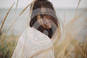 Portrait of beautiful stylish woman with windy hair in knitted sweater among wild grass at sea. Carefree calm moment. Fashionable