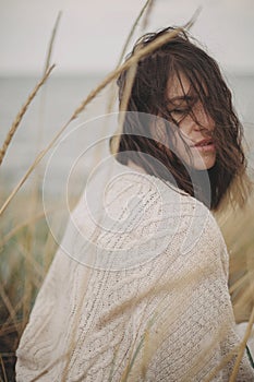 Portrait of beautiful stylish woman with windy hair in knitted sweater among wild grass at sea. Carefree calm moment. Fashionable