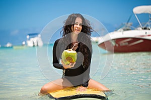 Portrait of a beautiful surfing girl with green coconut from palm tree sit on yellow surf longboard surfboard board.