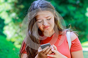 Portrait of a beautiful teen girl typing messages on the smart phone in a park with a green unfocused background