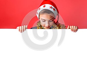 Portrait of a beautiful teen in Santa`s hat and with tinsel on her neck, posing behind a white panel isolated on a red background
